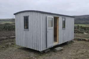 Shepherd’s hut exterior with light grey painted timber cladding, matching door and windows, set in a Highland landscape