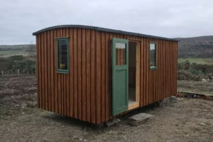 Shepherd’s hut exterior with dark stained timber cladding, green door and windows, set in a Highland landscape